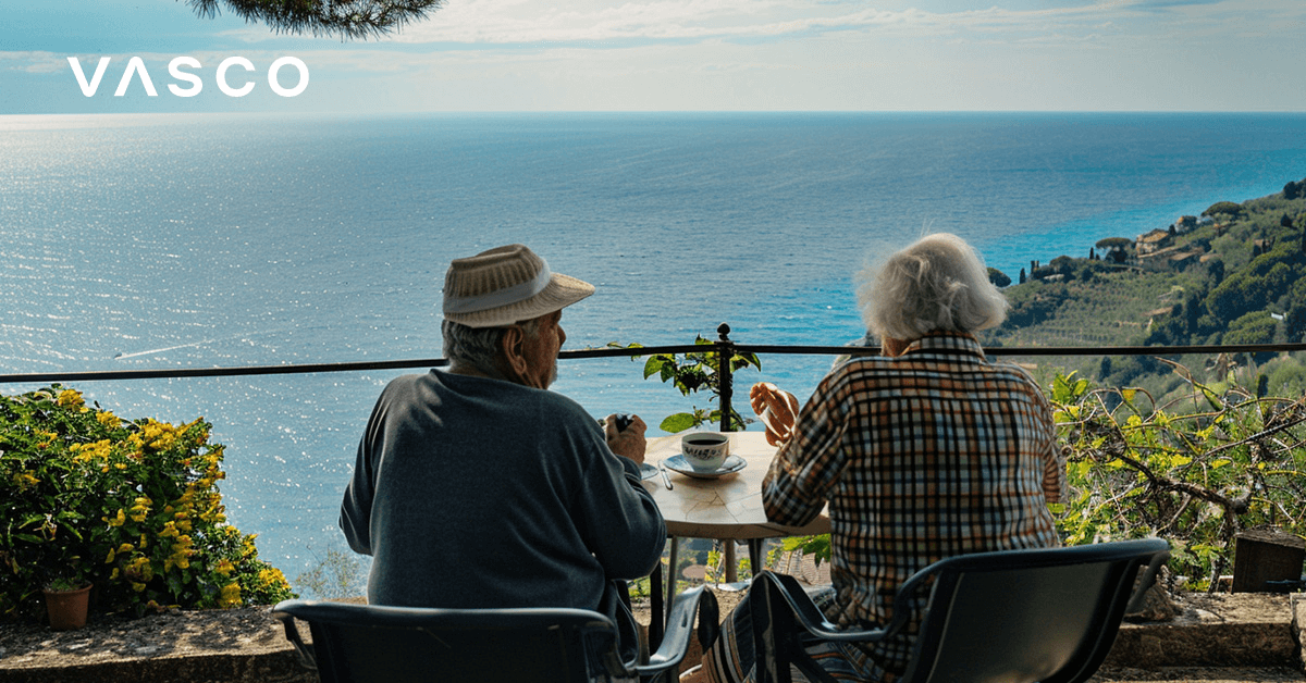 Un cuplu de seniori bea cafea pe un balcon cu vedere la mare.