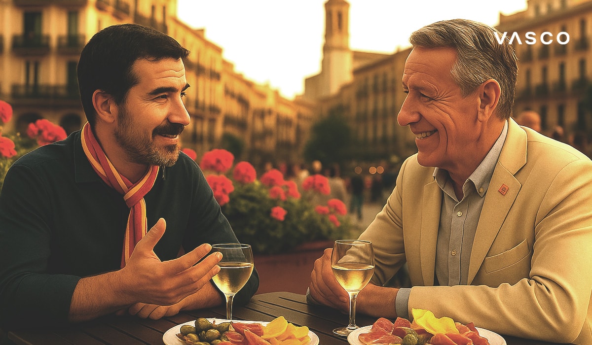 Doi bărbați savurând băuturi și tapas la o terasă dintr-o piață spaniolă, absorbiți de conversație, cu flori și arhitectură pe fundal.