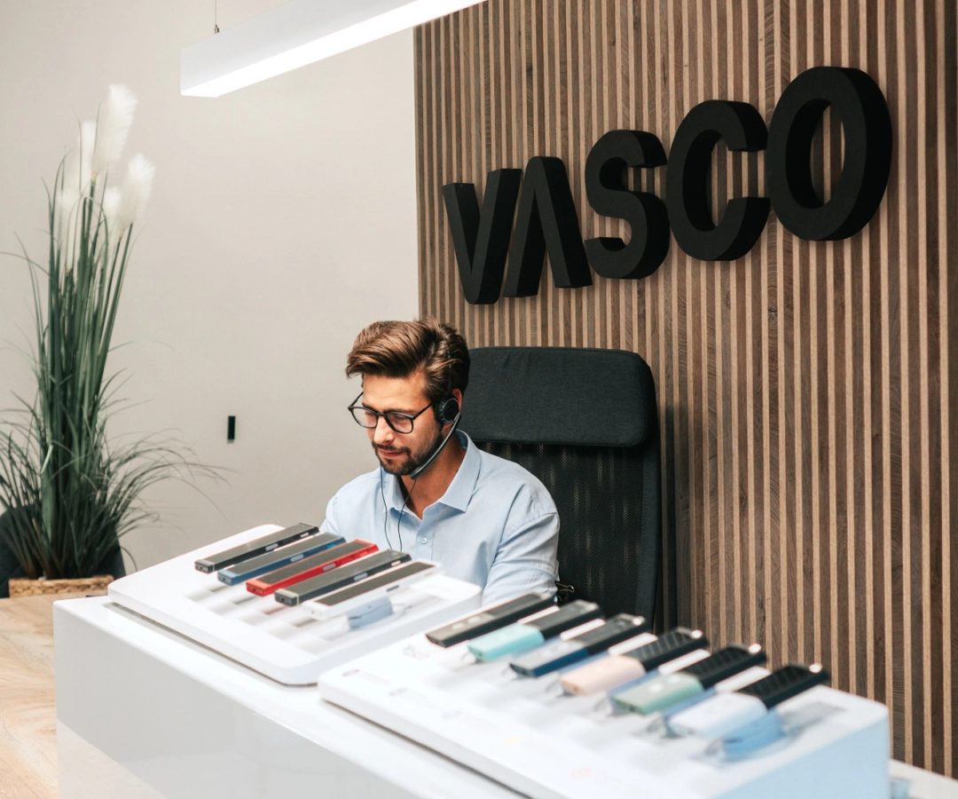 Un bărbat de la serviciul clienți stă la un birou care afișează mai multe dispozitive Vasco Translator în diferite culori, cu un semn mare „VASCO” pe peretele de lemn din spatele lui.