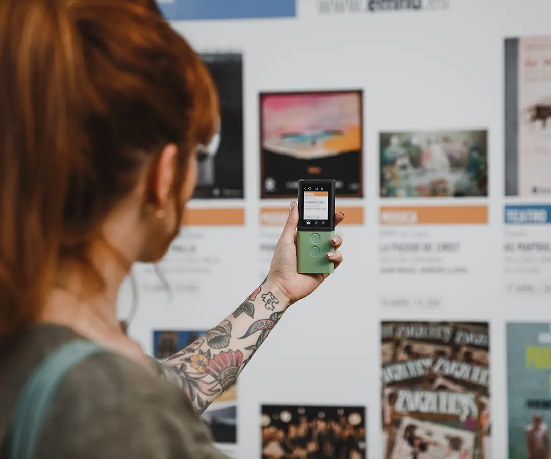O persoană roșcată cu un tatuaj floral pe braț ține în mână un dispozitiv digital verde, îndreptându-l spre un panou de afișaj cu diverse afișe și informații despre eveniment.