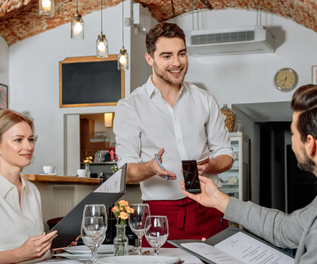 Un chelner zâmbitor stă la masa unui restaurant, în timp ce un client de sex masculin așezat ține un translator în mână. O clientă stă în apropiere, ținând în mână un meniu. Masa este aranjată cu pahare, farfurii și o mică vază cu flori.