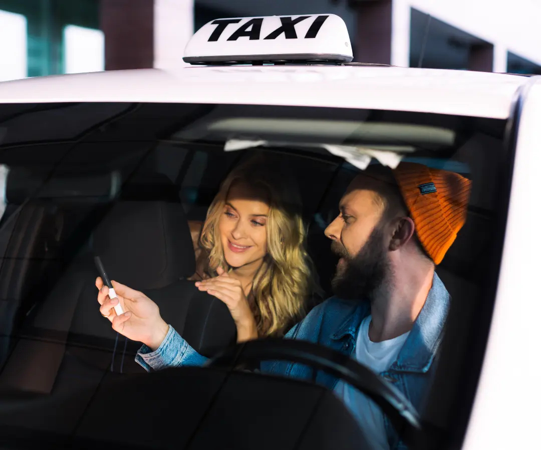 Un bărbat stă pe scaunul șoferului unui taxi și îi arată ceva unei femei de pe bancheta din spate pe un translator pe care îl ține în mână. Femeia zâmbește cu interes. Un semn de taxi este vizibil pe acoperișul mașinii.