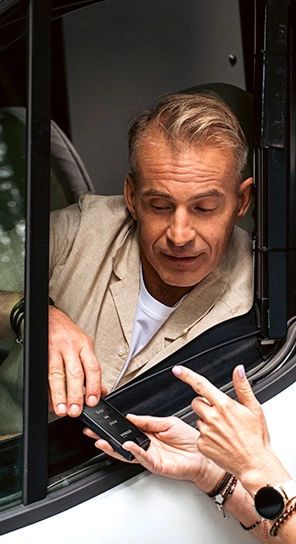 Un bărbat aflat la volan îi vorbește dispozitivului Vasco Q1, pe care cineva i-l oferă prin geam.