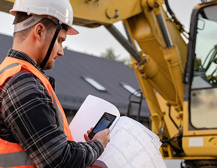 Un bărbat purtând cască și haine de lucru fotografiază planuri de construcție folosind Vasco Translator V4, cu un excavator vizibil în fundal.