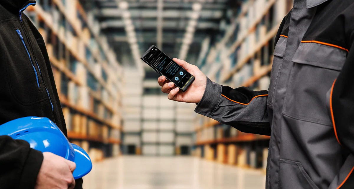 Două persoane stau față în față; una ține o cască, iar cealaltă ține un Vasco Translator V4. Ecranul dispozitivului afișează interfața activă de traducere vocală.