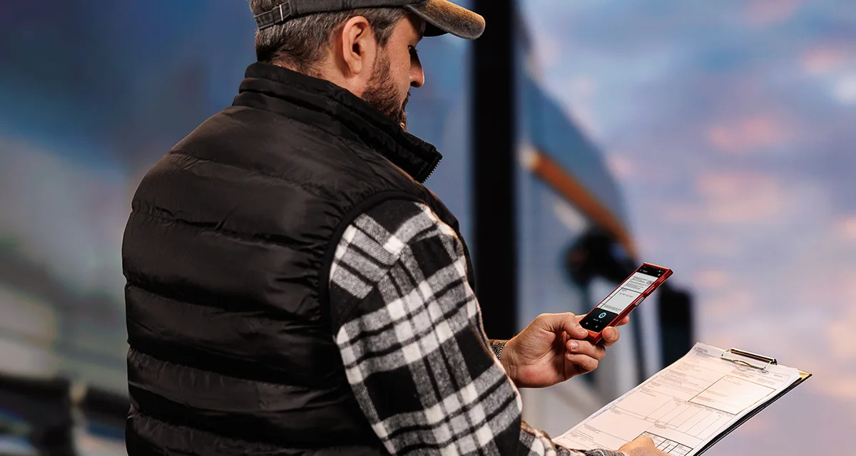 Un șofer de camion stă în fața camionului, ținând documente într-o mână și un Vasco Translator V4 în cealaltă. Ecranul dispozitivului afișează funcția de traducere prin fotografie.