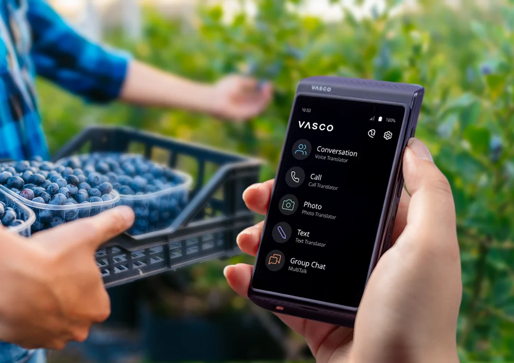 Vasco Translator Q1 în prim-plan, cu tufe de afine în fundal și o persoană care ține o lădiță cu afine recoltate.