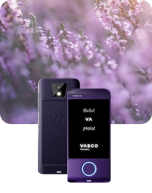 O imagine de prezentare a produsului în care apare un dispozitiv Vasco Translator de culoare violet, surprins din două unghiuri, pe un fundal din țesătură închisă la culoare și cu textură. Dispozitivul din stânga prezintă partea din spate, punând în evidență obiectivul camerei cu bliț și logo-ul „VA” în relief. Dispozitivul din dreapta prezintă vederea frontală cu ecranul iluminat, afișând saluturile „Hello!”, „VA”, „¡Hola!” și logo-ul „VASCO Translator”. Un buton circular din partea de jos a dispozitivului este conturat cu o lumină albastră strălucitoare. Compoziția este așezată pe un fundal visător, estompat, cu flori de erică violetă scăldate în lumina blândă a soarelui.
