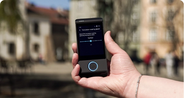 Un prim-plan al mâinii unei persoane care ține un traducător electronic Vasco de culoare neagră, pe fundalul estompat al unei piețe din oraș, cu clădiri istorice. Ecranul dispozitivului afișează setările pentru „Viteza de citire a traducerii”, cu un glisor setat pe „Normal” și o notă care spune: „Reglați viteza de citire a traducerii mutând glisorul.” Persoana care ține dispozitivul poartă o brățară argintie. Lumina puternică a zilei pune în evidență interfața ecranului, făcând setările ușor de văzut. 